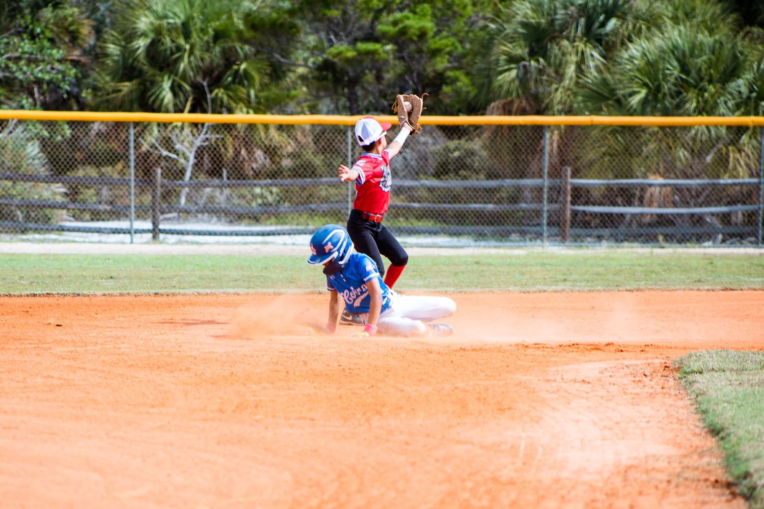 Youth baseball players diving for the ball on an orange clay infield during a competitive game.