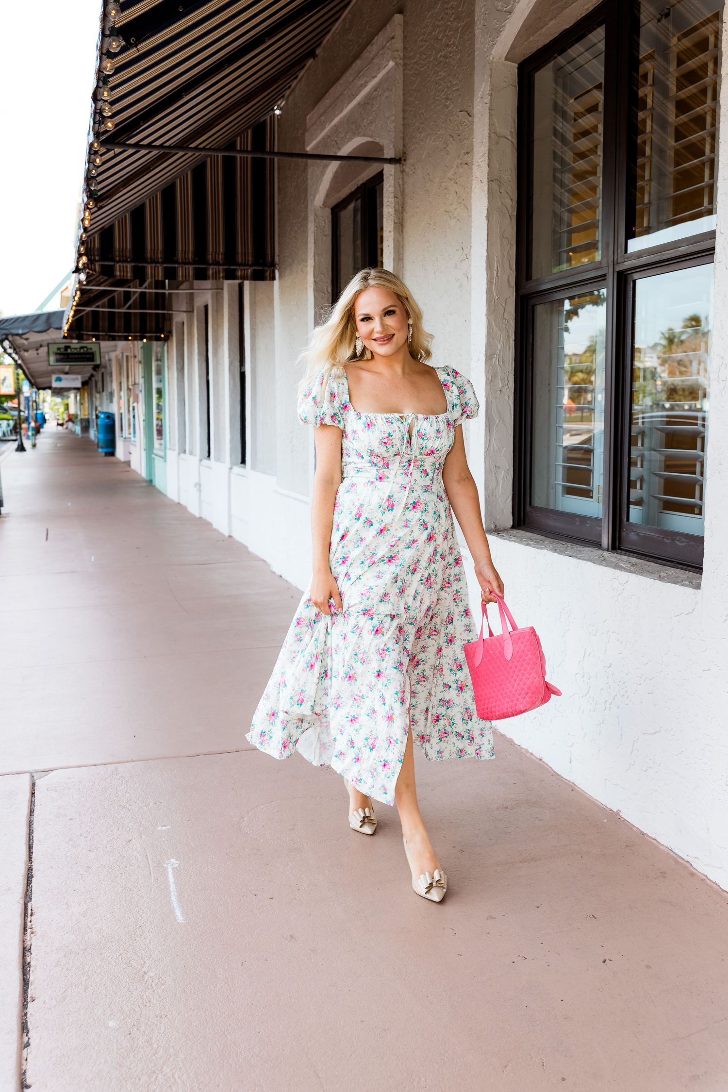 A woman in a flowing floral dress walks down a sidewalk carrying a pink handbag.