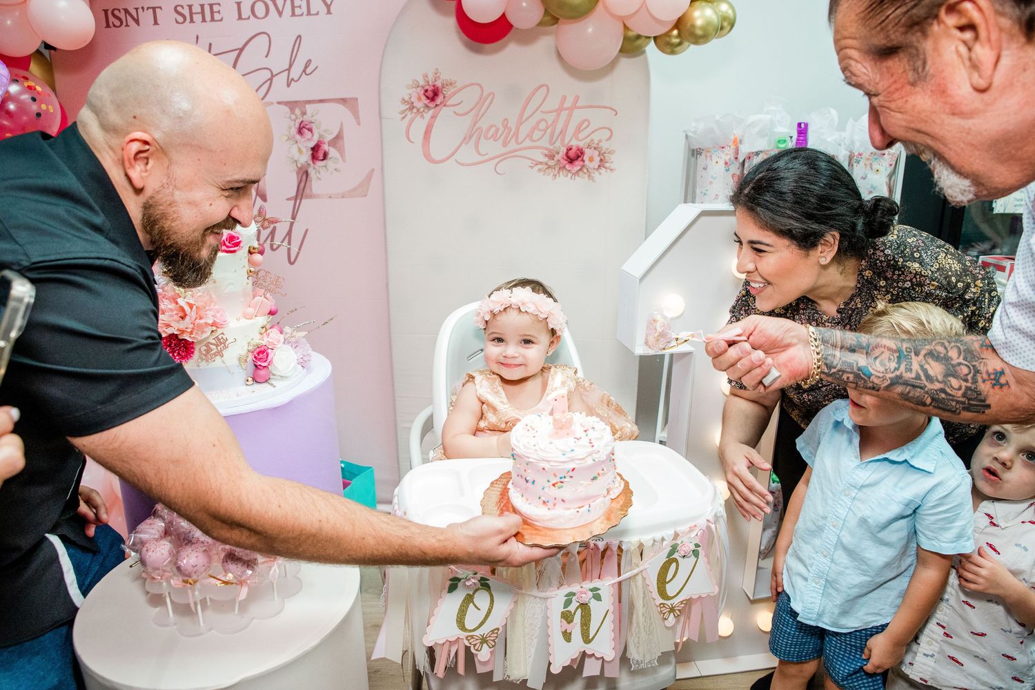 Baby girl celebrates first birthday in high chair with pink cake, surrounded by family at a decorated party.