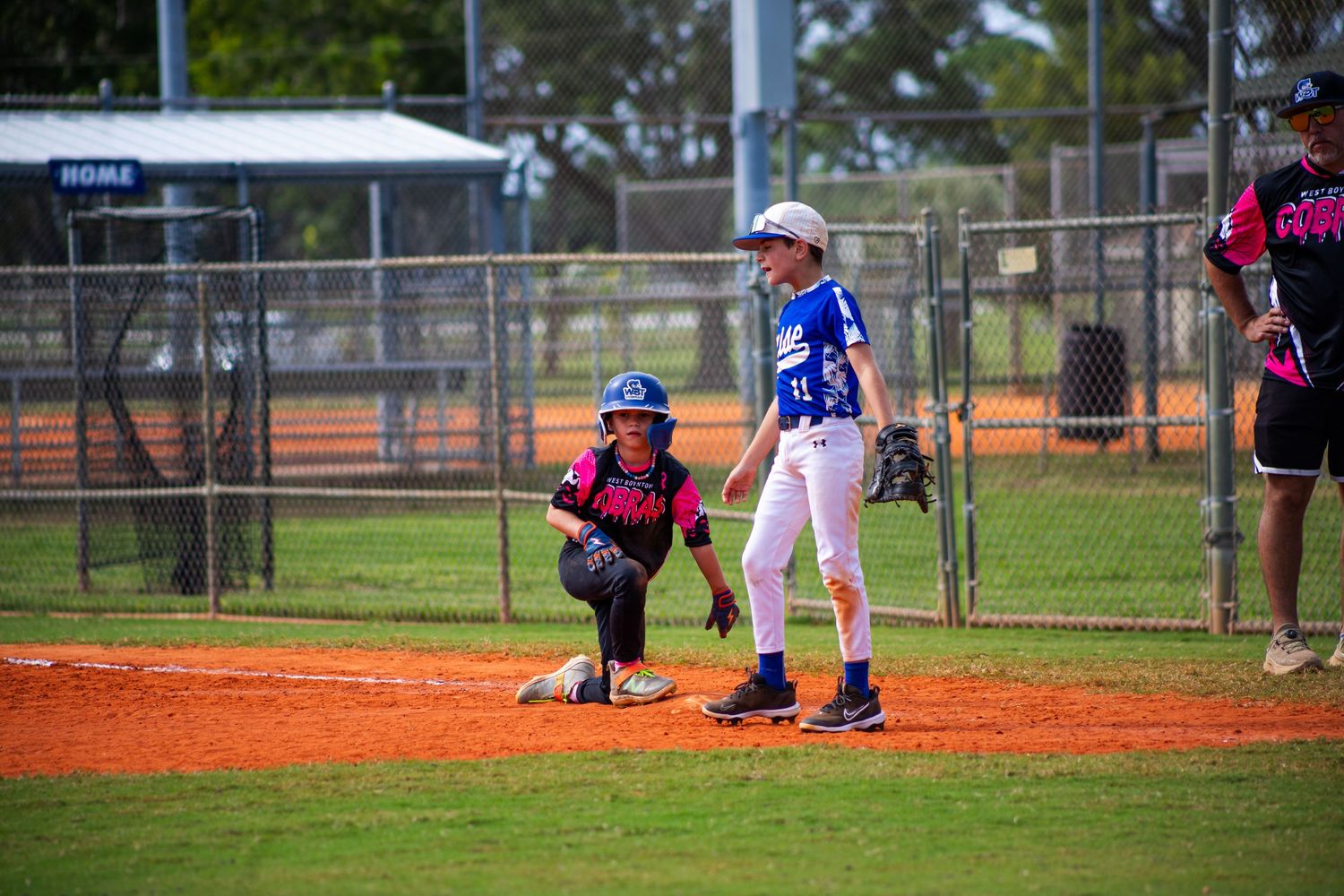 Youth baseball game in progress with batter at plate and catcher in position on dirt diamond with green grass outfield.