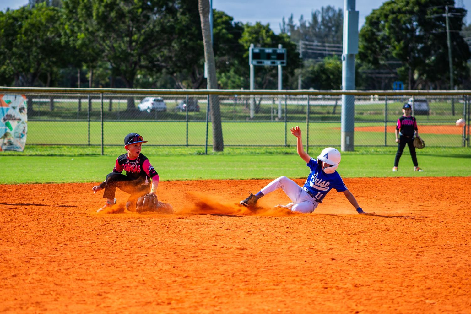A baseball player slides into base during a game on an orange clay infield while another player reaches to make the tag.