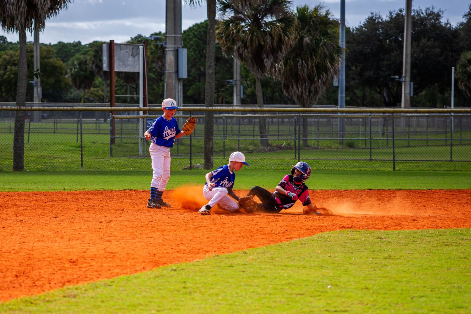 Baseball players slide into base during an intense play on a bright orange dirt infield at a baseball field.