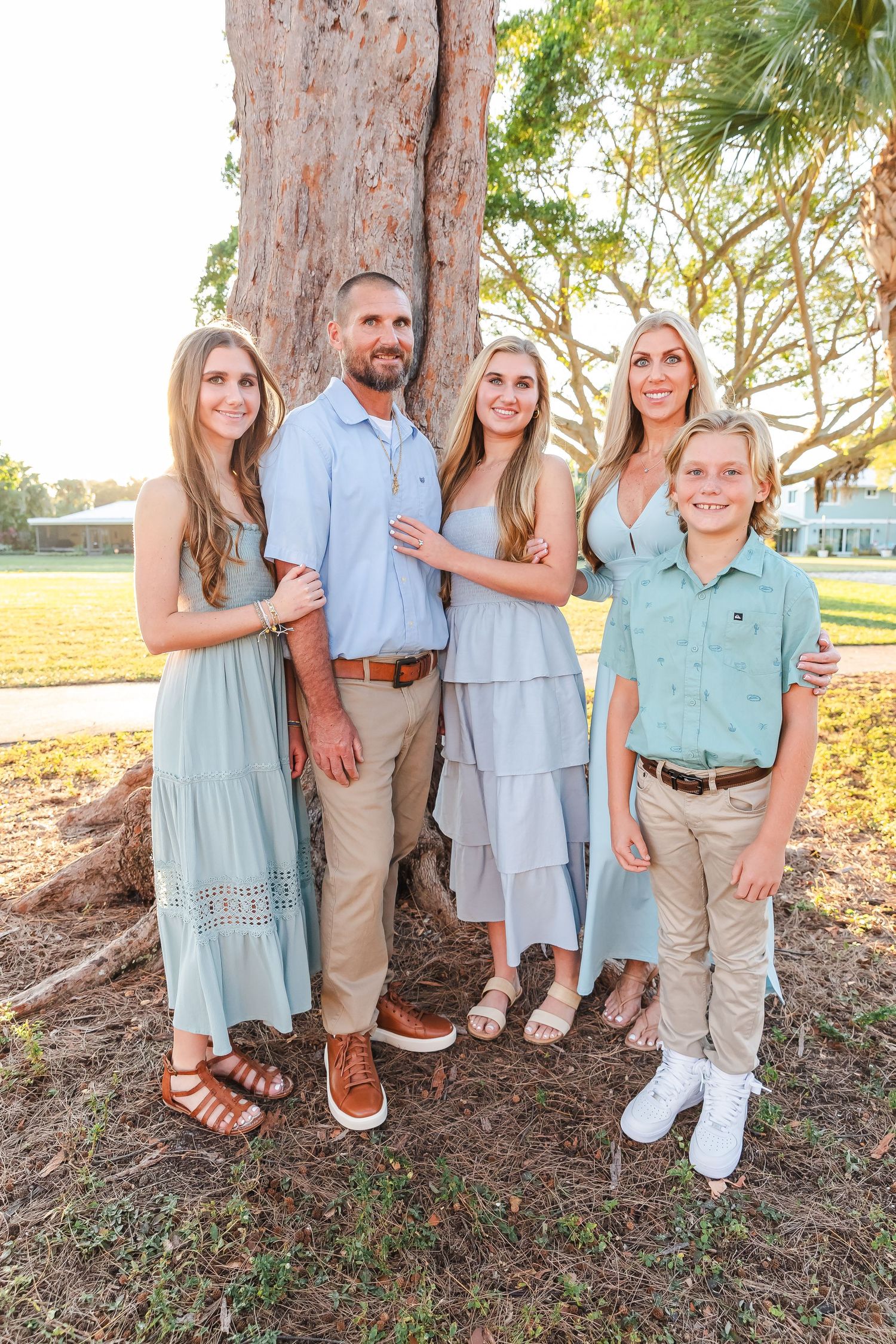 Family group dressed in coordinating pastel blue and khaki outfits poses near palm trees outdoors.