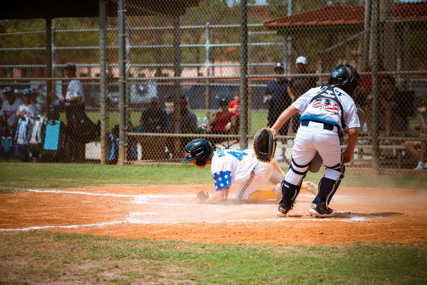 A baseball player slides into home plate while the catcher awaits with glove ready during a game on a dirt field.