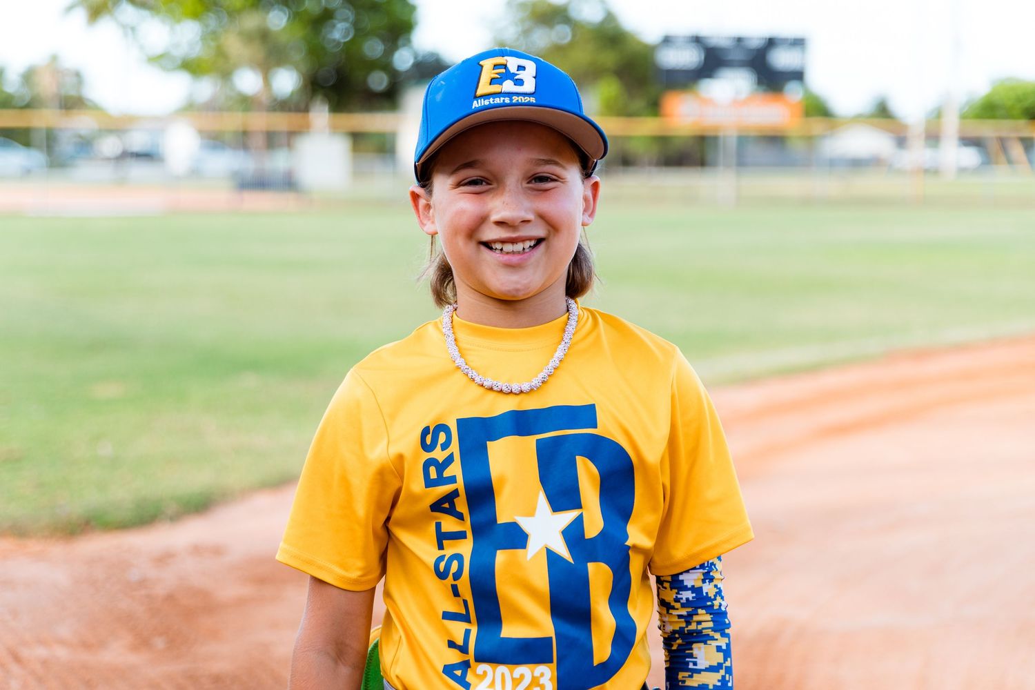 Youth baseball player wearing a yellow jersey and blue cap stands on baseball field with a warm smile.