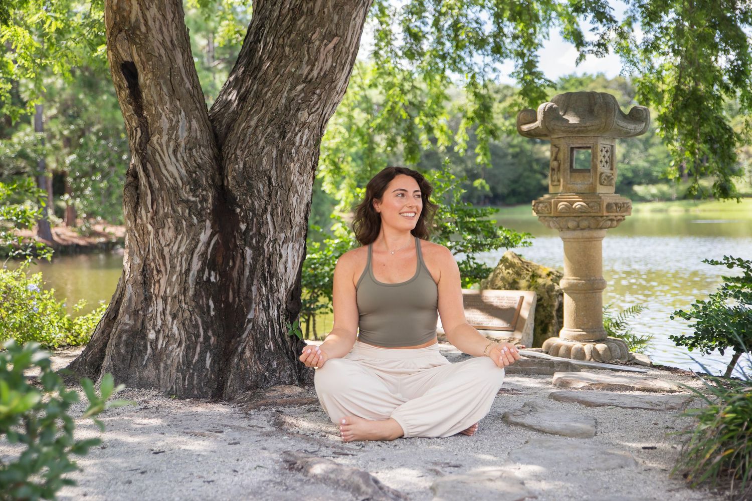 A person meditating in lotus pose beneath a large tree near an ornamental stone lantern and pond in a peaceful garden setting.