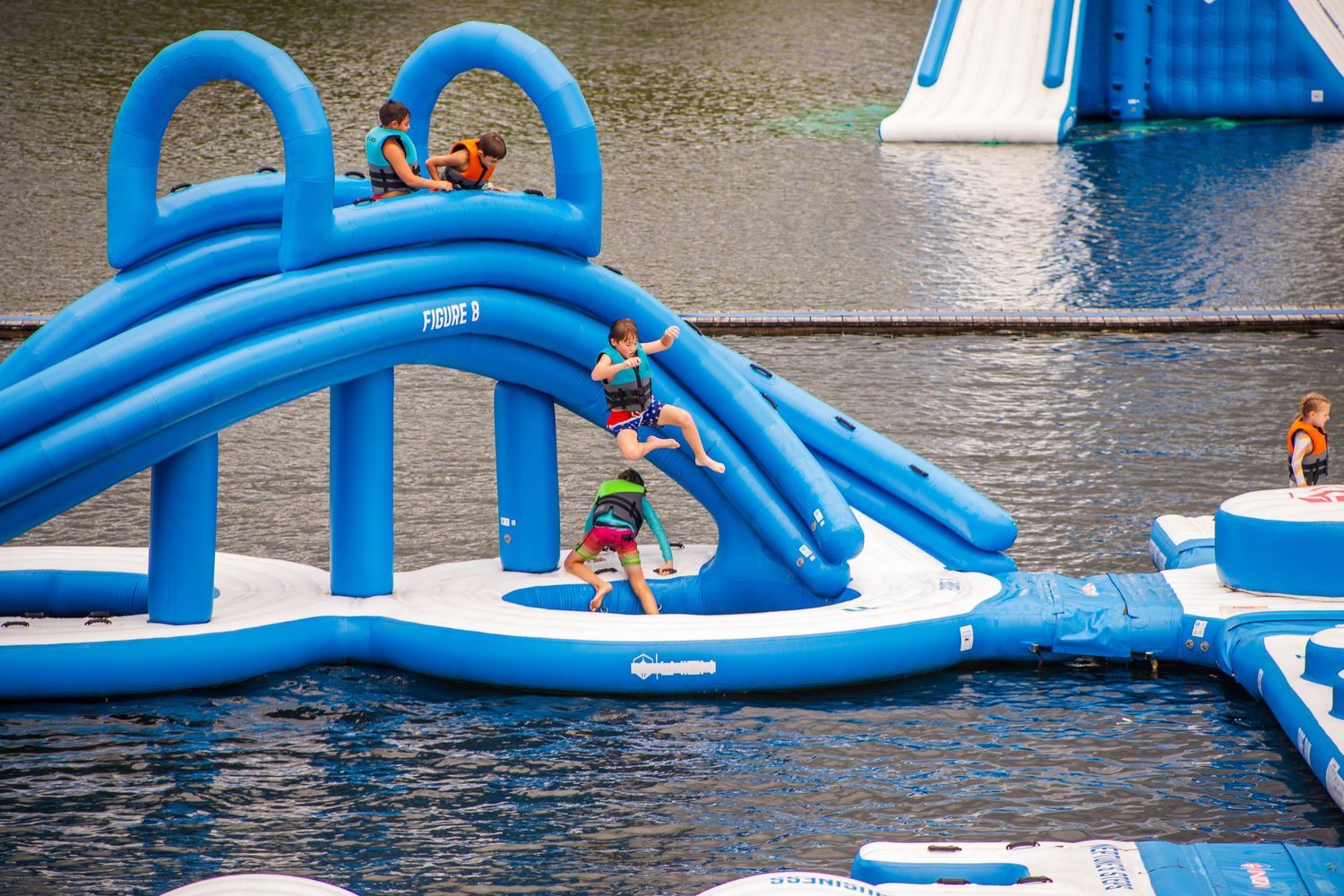 Blue inflatable water obstacle course on a lake with people enjoying activities on a sunny day.