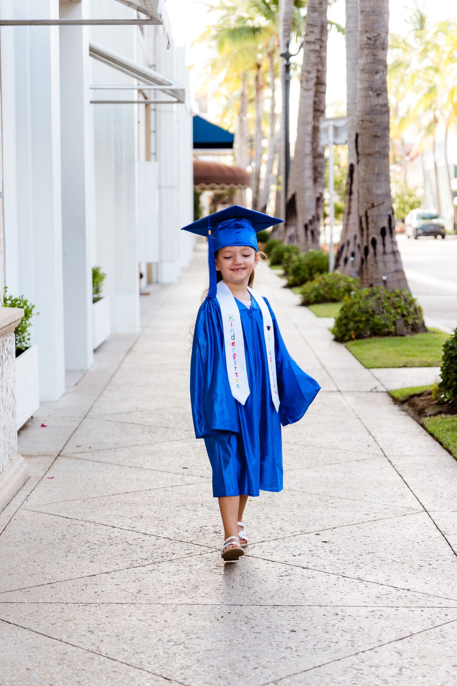 Small graduate in blue cap and gown walks along palm tree-lined sidewalk wearing white tie.