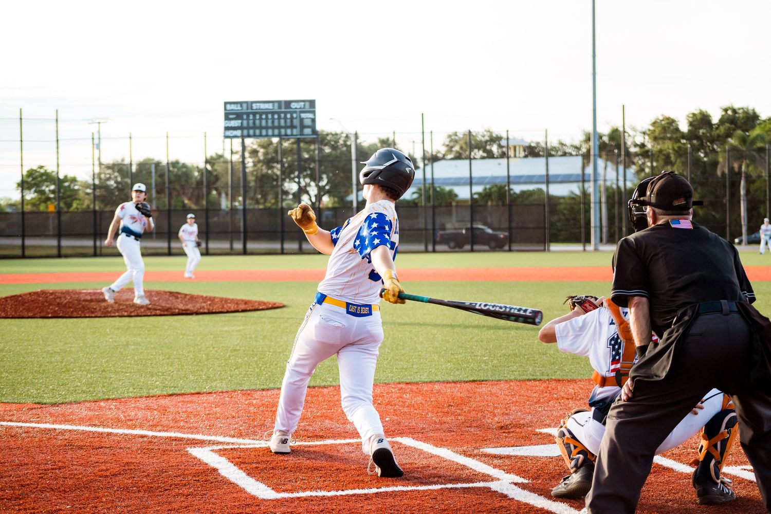 Baseball player at bat in white and blue uniform stands ready at home plate while umpire watches nearby during game.