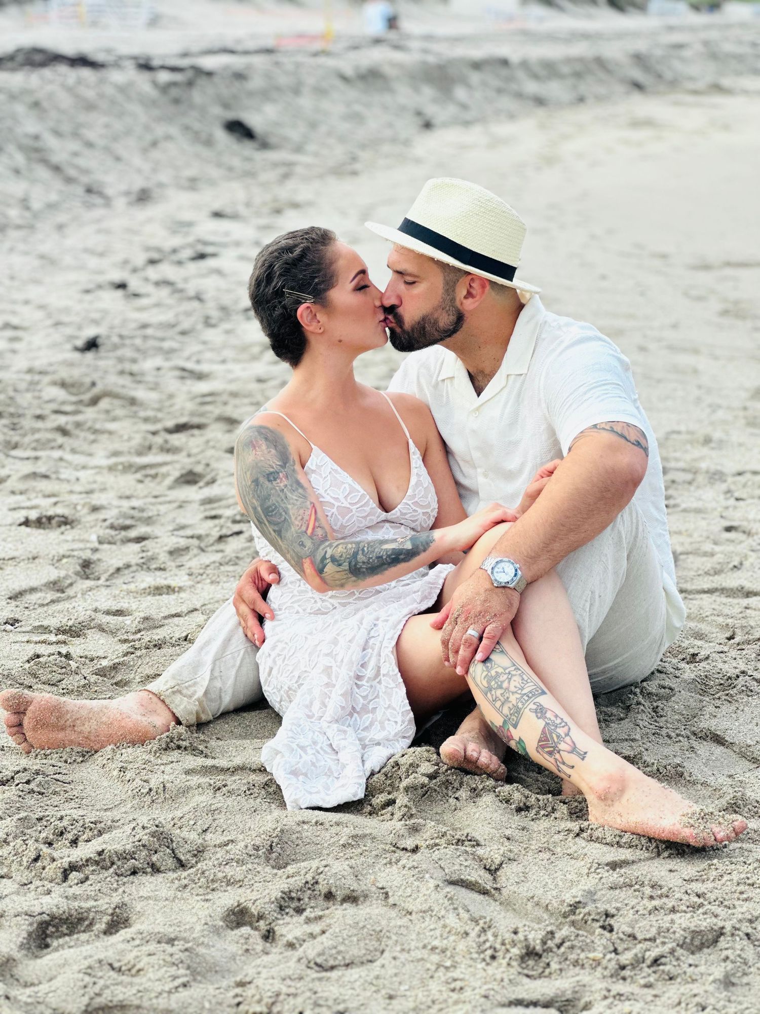 A couple shares an intimate moment while sitting on the beach sand wearing white summer attire.