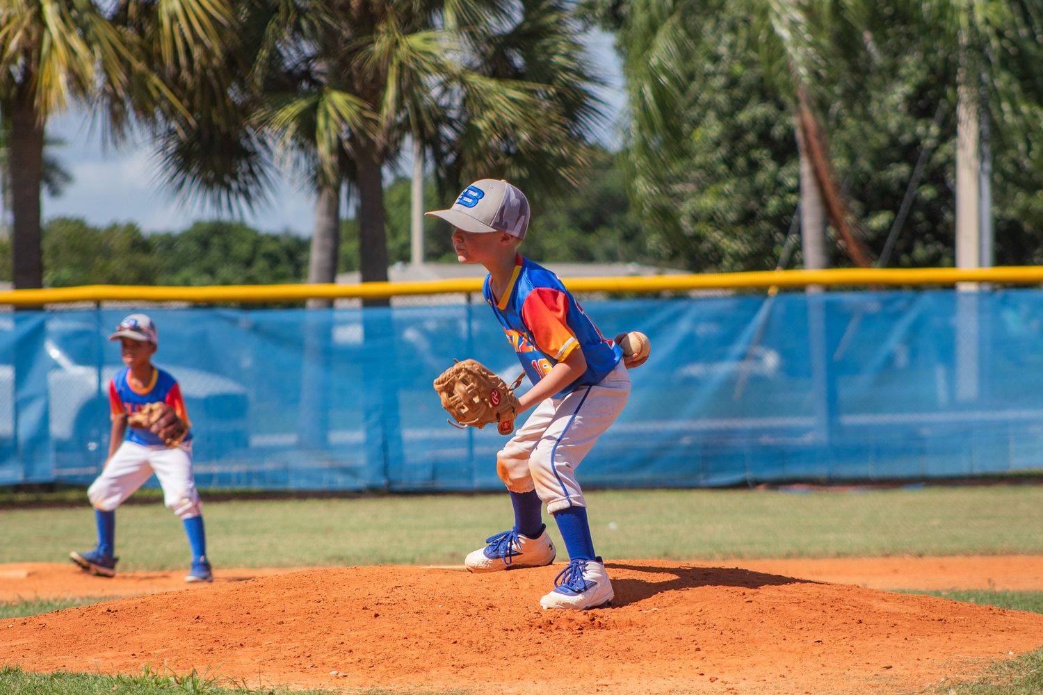 A baseball pitcher on the mound delivers a pitch during a youth game with blue outfield wall visible.