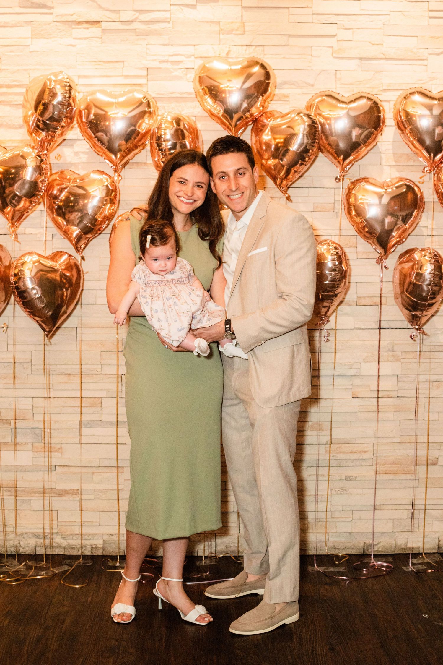 Couple in green dress and beige suit holding baby with rose gold heart balloons backdrop.
