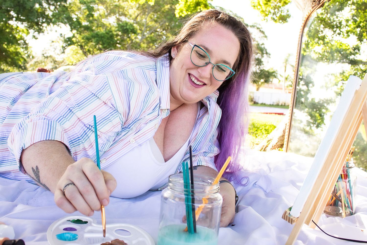 Artist painting outdoors on white blanket with colorful art supplies scattered around.