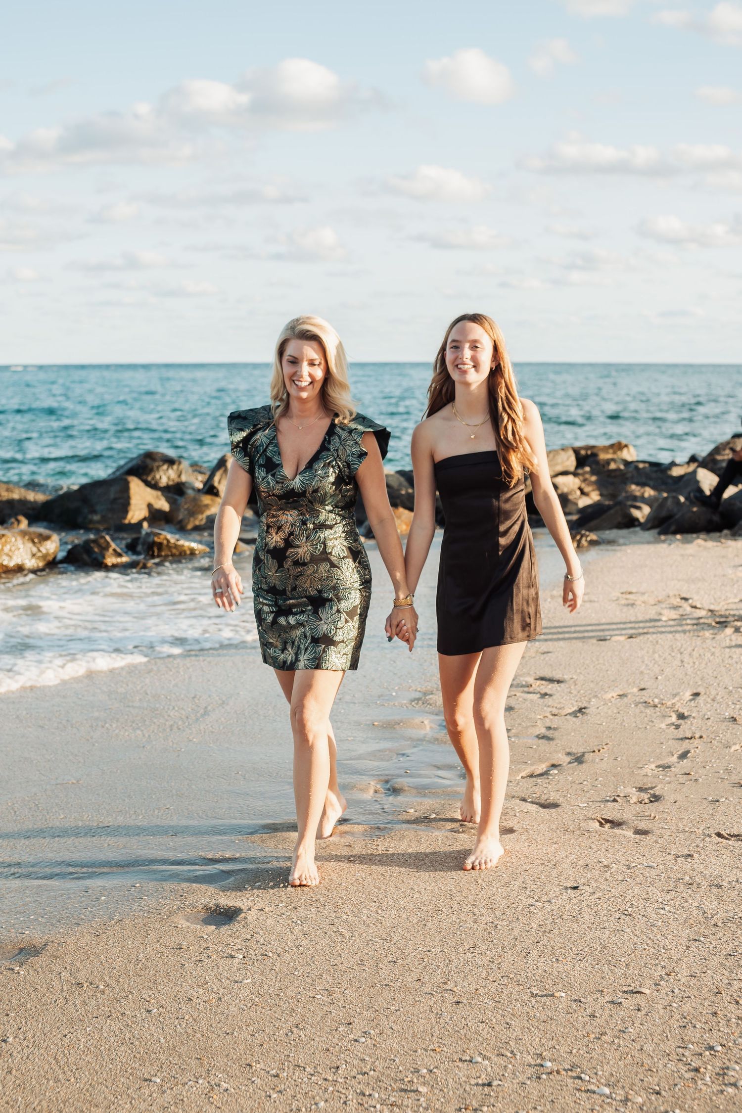 Two people walking hand in hand on a tropical beach with rocks and turquoise water in the background.