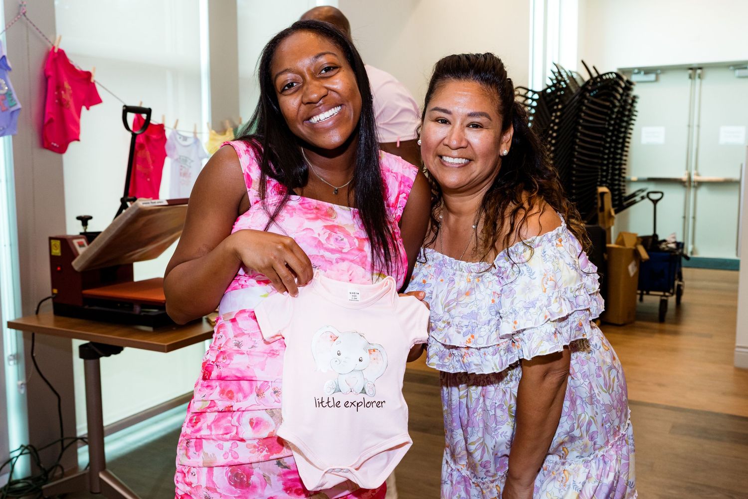 Two friends show off a pink baby onesie at a baby shower celebration.