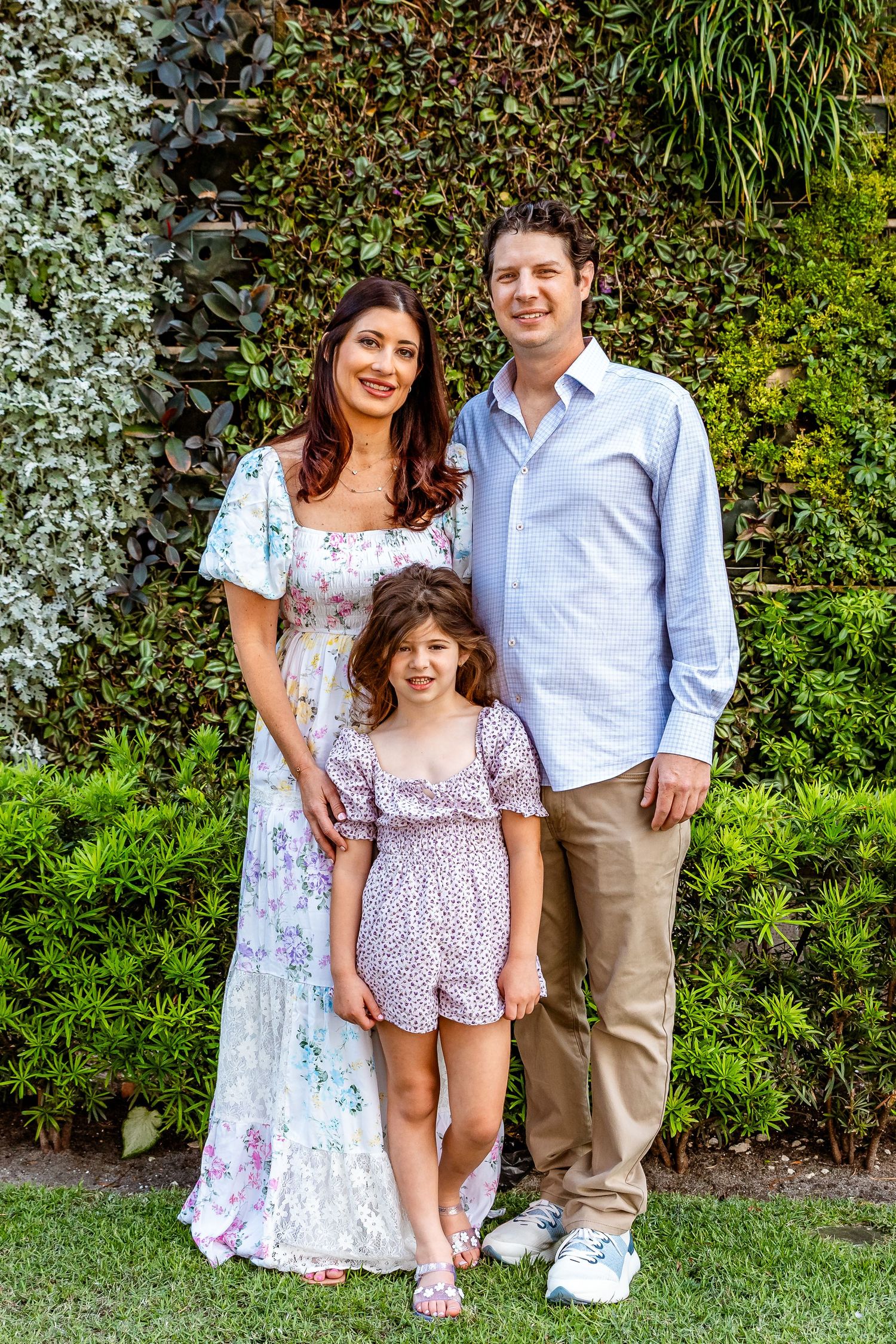 Family of three posing in front of a green plant wall, mother in floral dress with father and daughter.