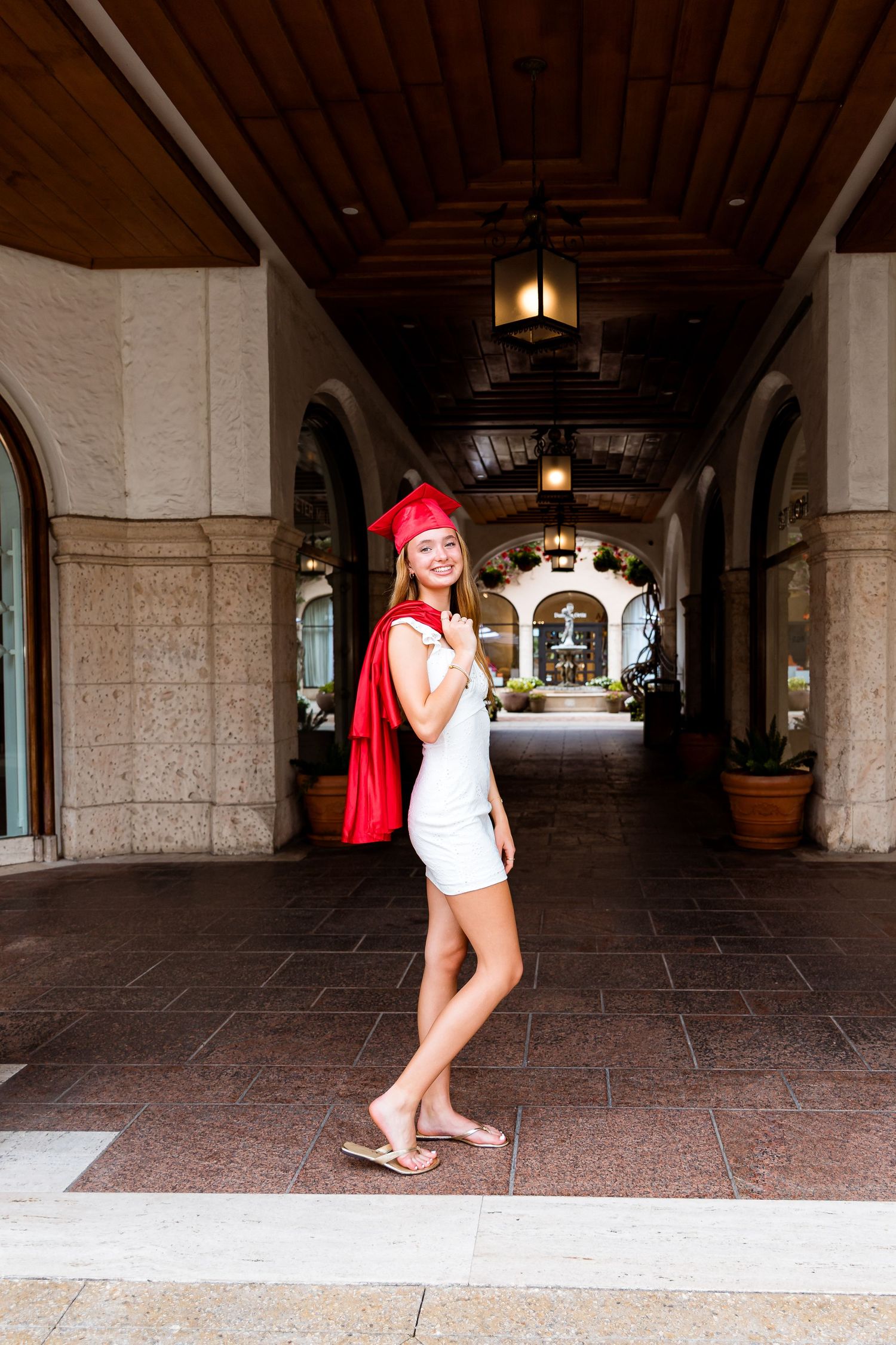 A woman in a white dress and bright red cape stands in an elegant arched hallway with hanging lanterns.
