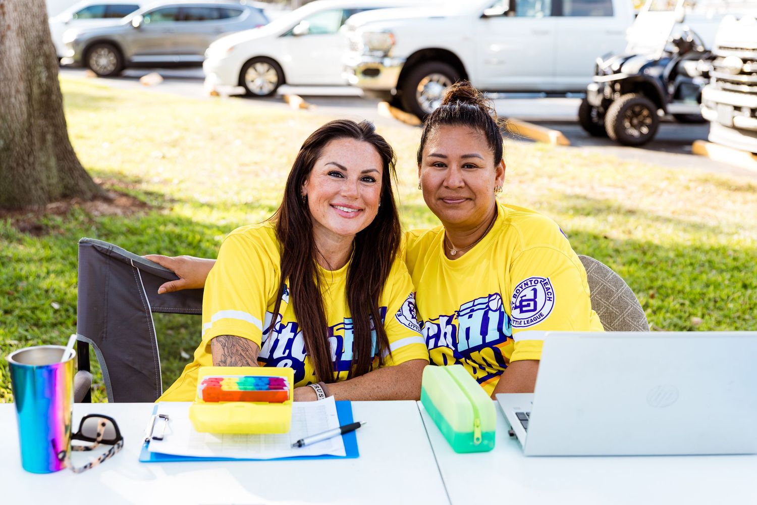 Two people wearing yellow shirts sitting at an outdoor table with a laptop and drinks.