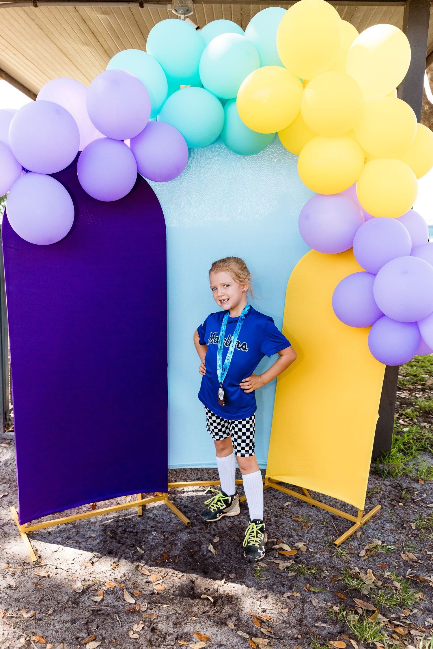 A colorful balloon arch backdrop in purple, blue and yellow creates a festive photo backdrop against an outdoor setting.