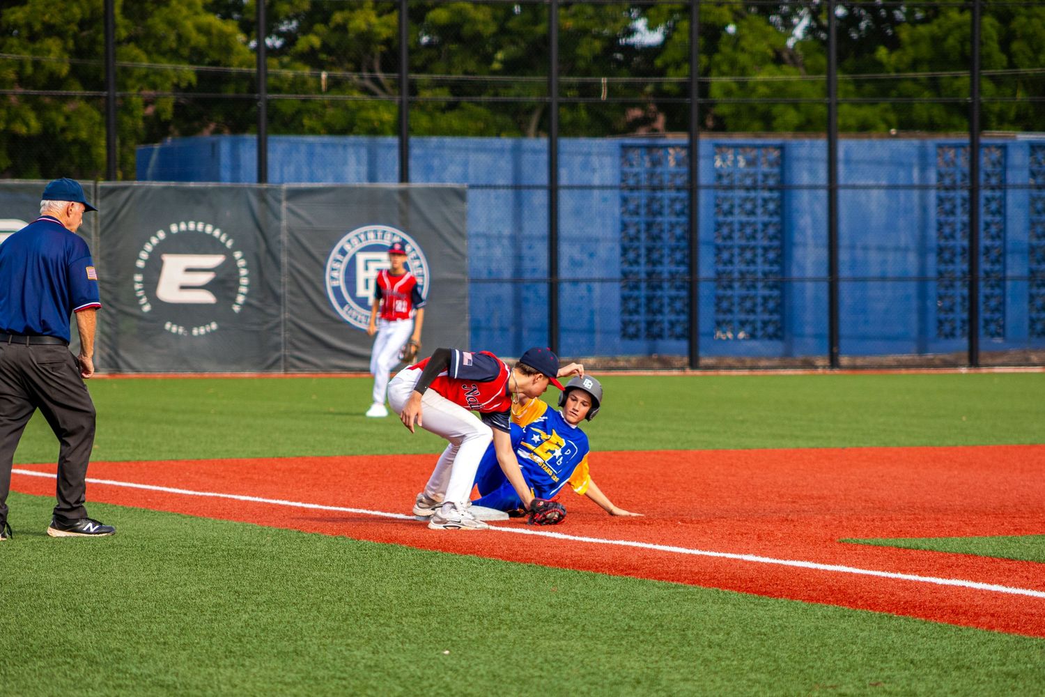 Baseball player sliding into base during a competitive game on a red dirt field with blue outfield wall.