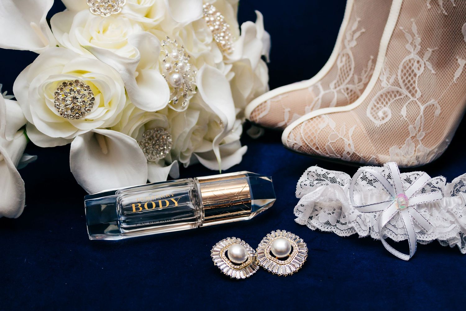 Elegant wedding details with white roses, pearls, jewelry and high heel shoes arranged on reflective surface.