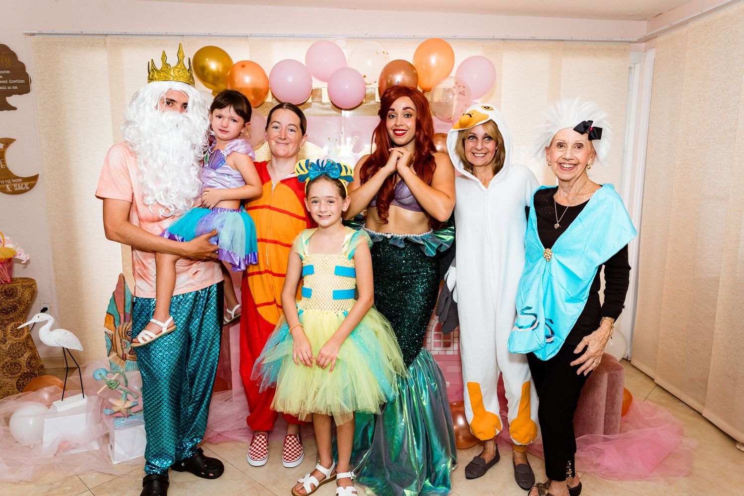 A group in colorful mermaid and sea-themed costumes pose together at a party with pink and gold balloons.
