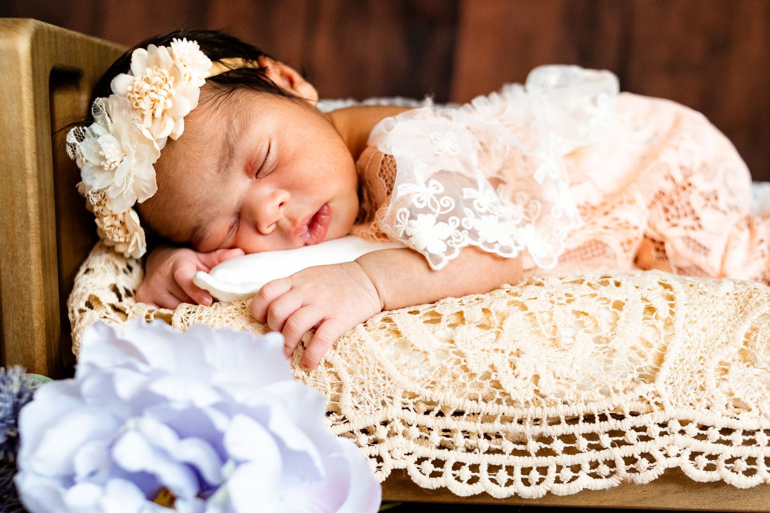 Sleeping newborn baby girl in lace dress and floral headband resting in a wooden crib with decorative doily.