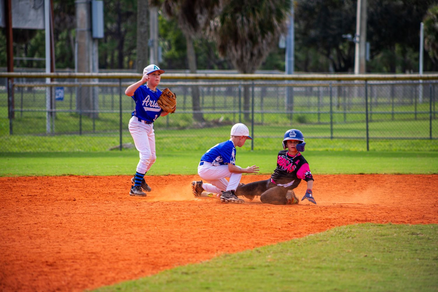 Baseball player slides into base during intense game action on dirt infield while opponent attempts to make the tag.