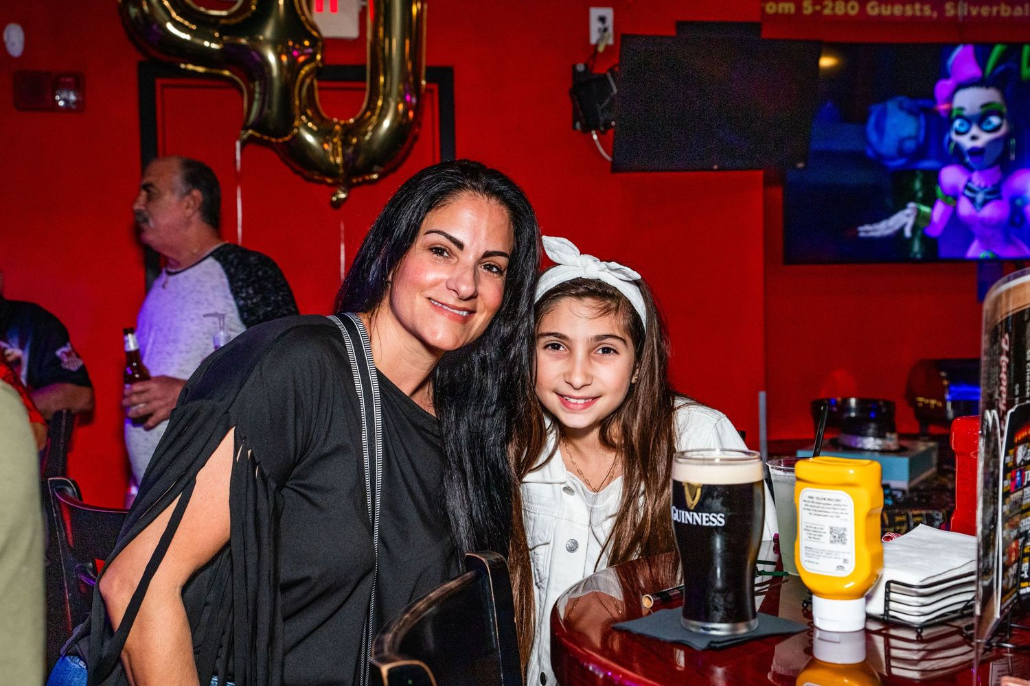 Two women posing together in a bar or restaurant setting, with colorful lighting and decorations in the background.