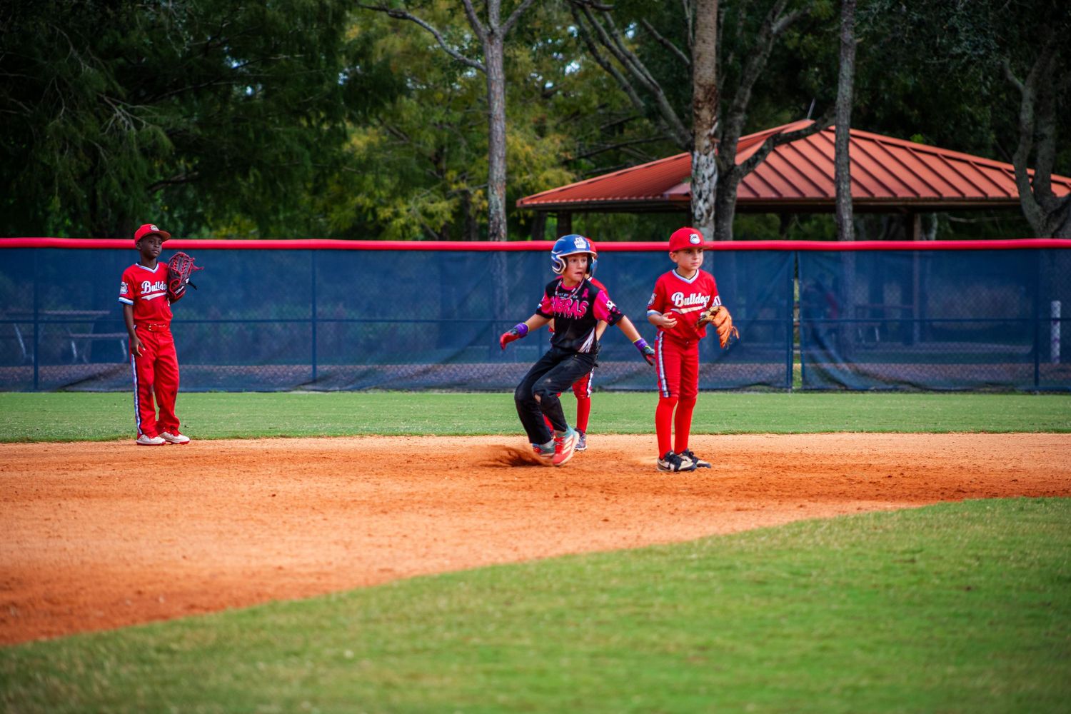Baseball players on field during game with red fencing and green trees in background.