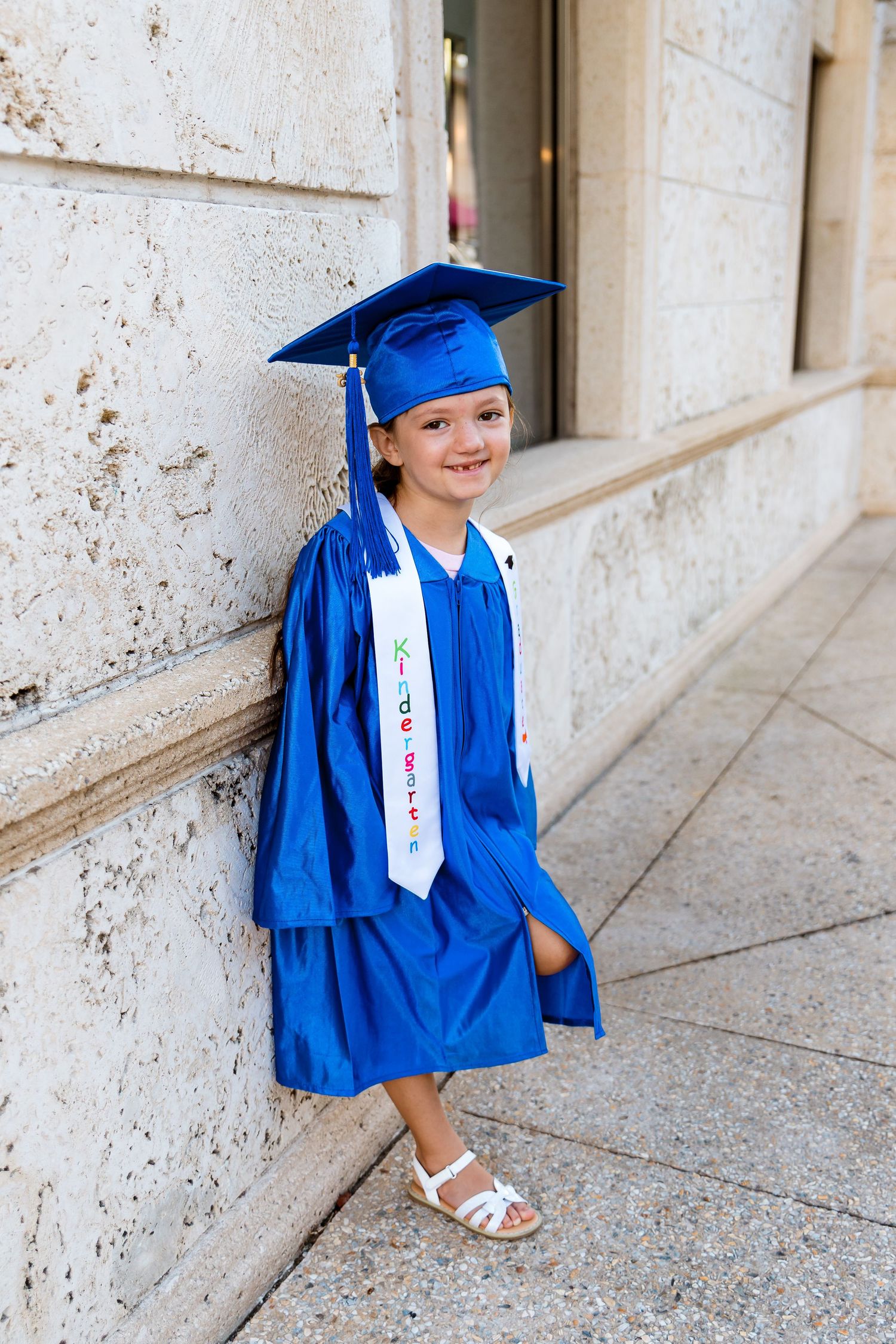 A young graduate in blue cap and gown poses while sitting against a stone wall during graduation photos.