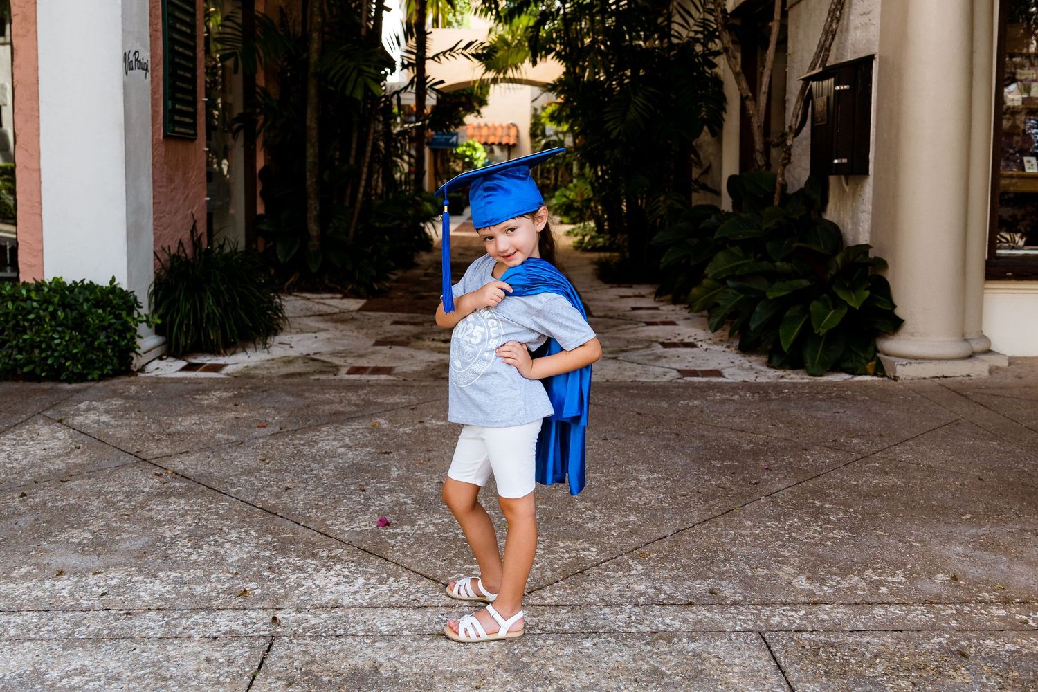 A figure wearing a bright blue graduation cap and gown stands on a walkway between white pillars and greenery.