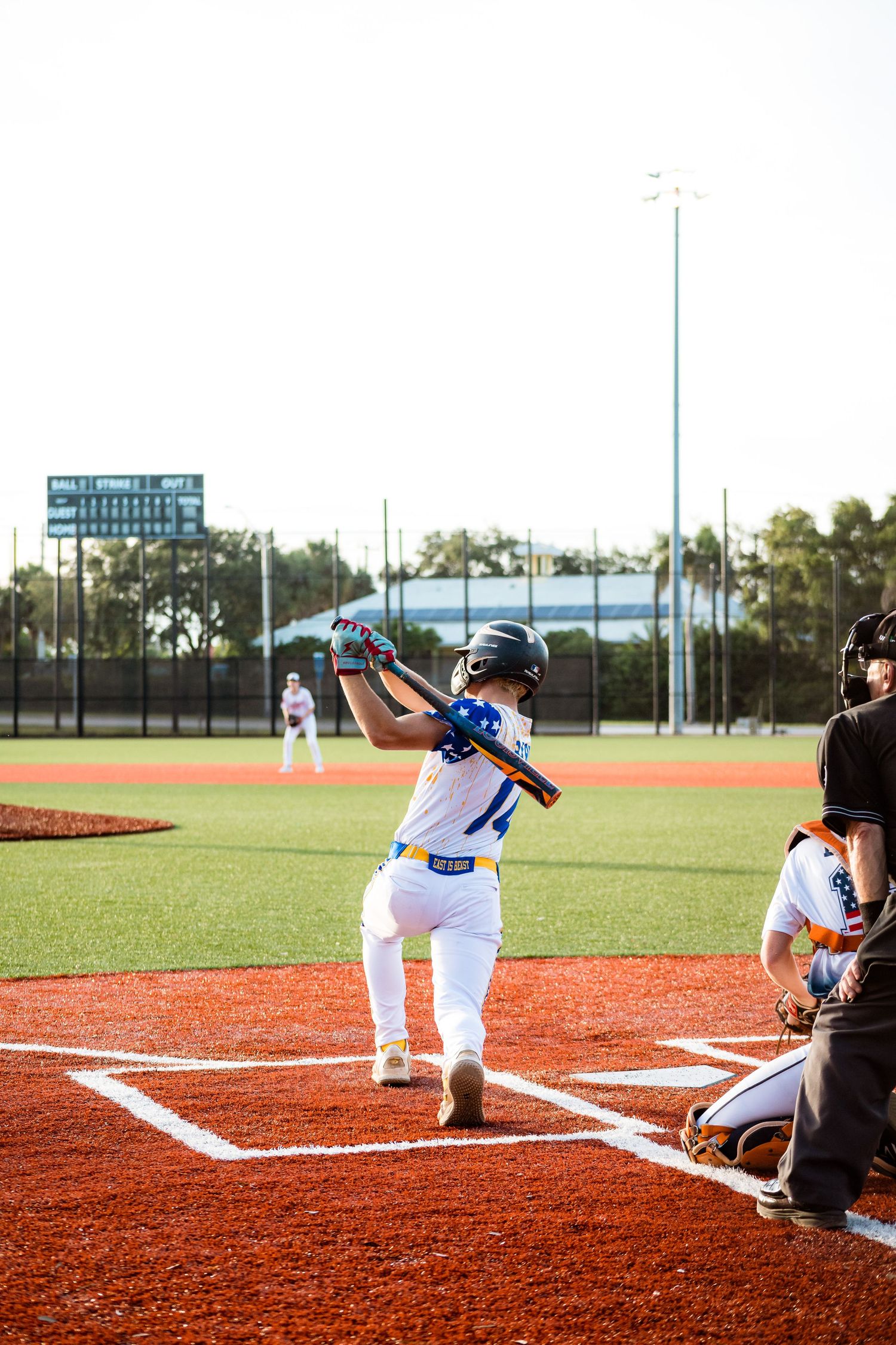 Youth baseball player in white uniform stands at home plate in batting stance during daytime game on baseball field.