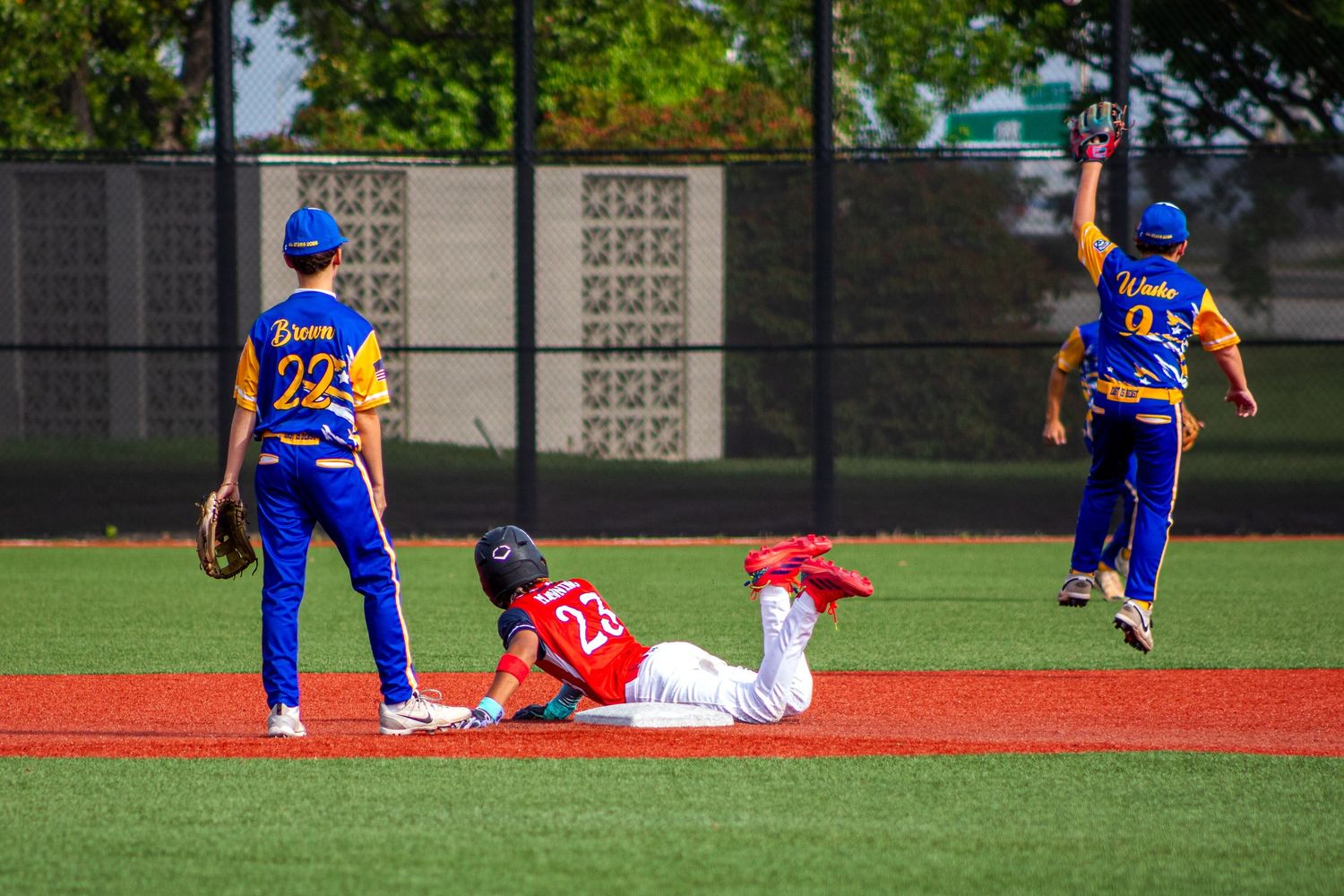 Youth baseball player slides into second base as fielders in blue and yellow uniforms react during a sunny game.