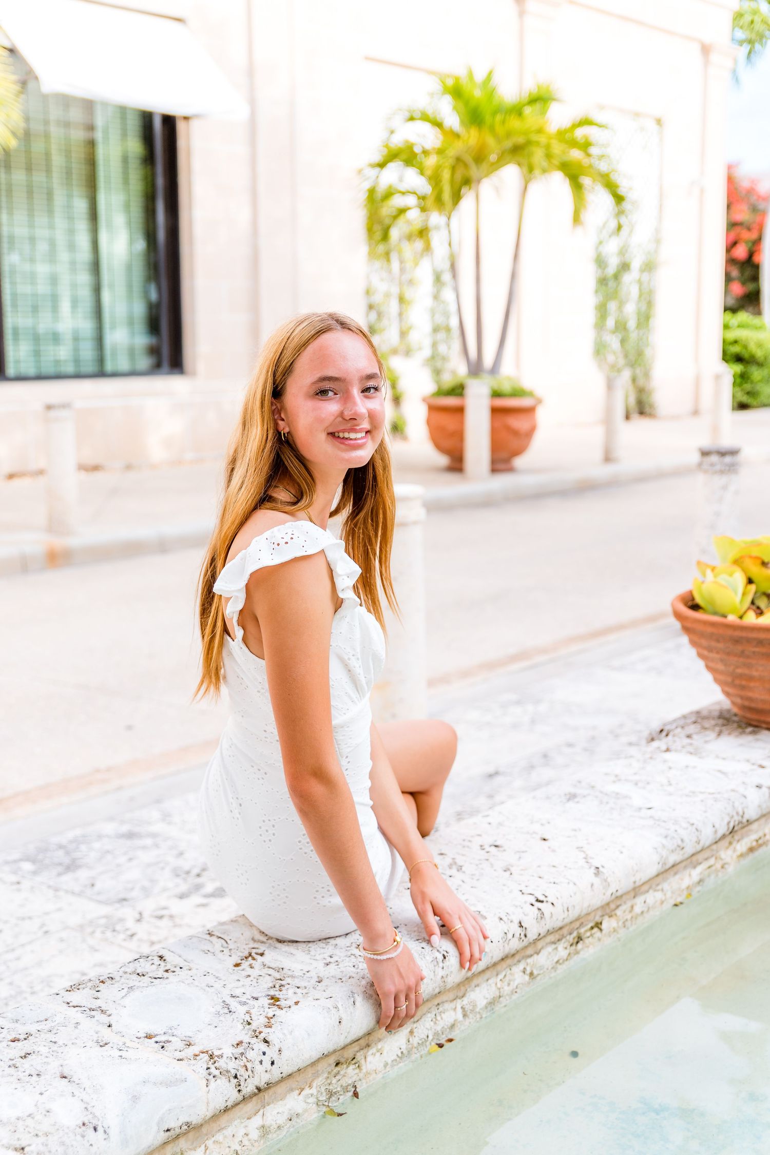 Person wearing white dress sitting on edge of fountain with tropical plants in background.