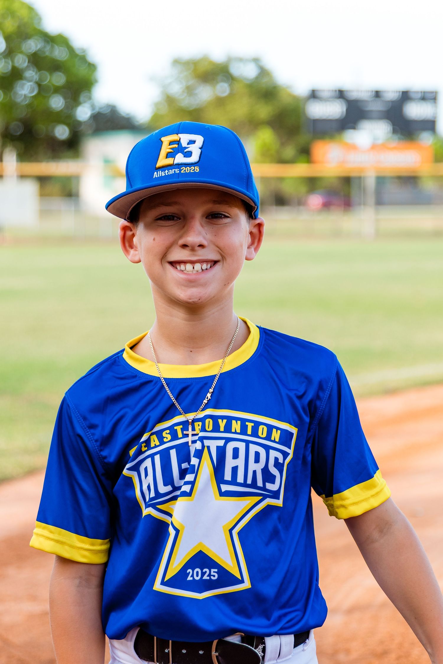Young baseball player wearing a blue and yellow East Covina All-Stars uniform and baseball cap on a dirt field.