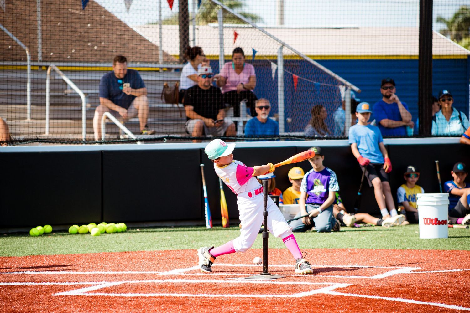 Youth softball player in purple uniform swings bat at tee ball during practice with spectators watching from bleachers.