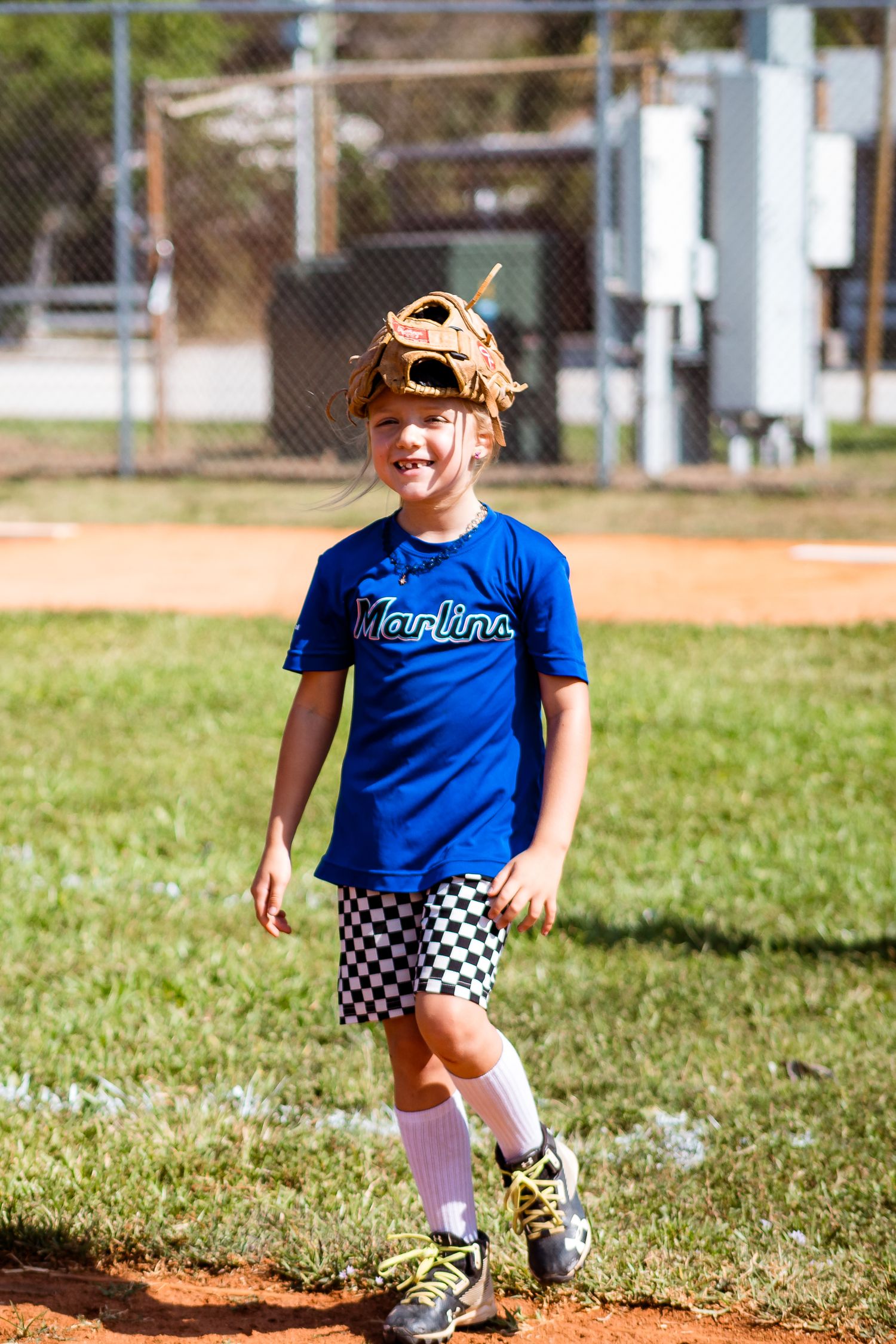 Young baseball player wearing a blue jersey and checkered shorts stands on a dirt baseball field in athletic gear.