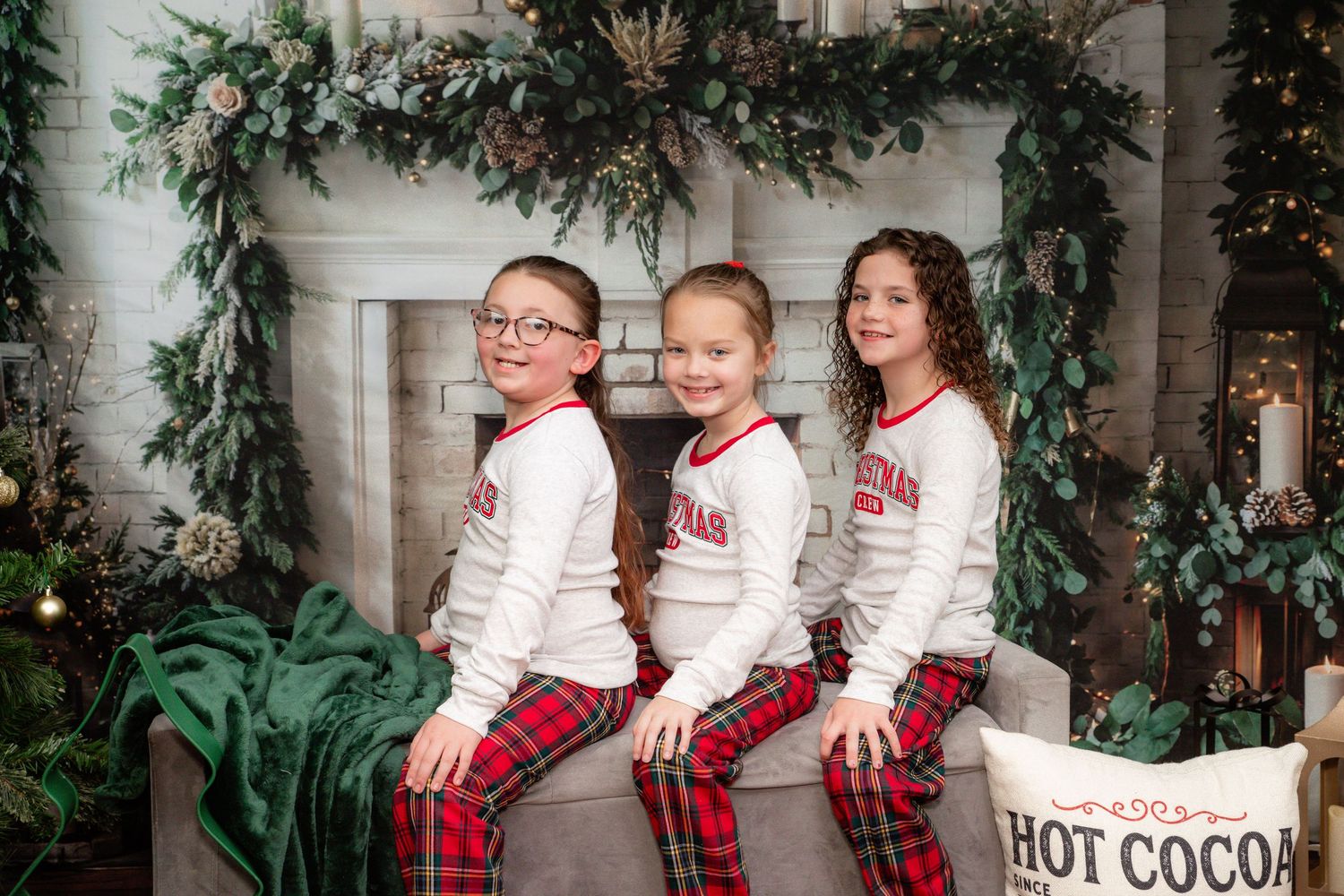 A group in matching plaid pajamas and white sweaters sits on stairs decorated with holiday greenery garlands.