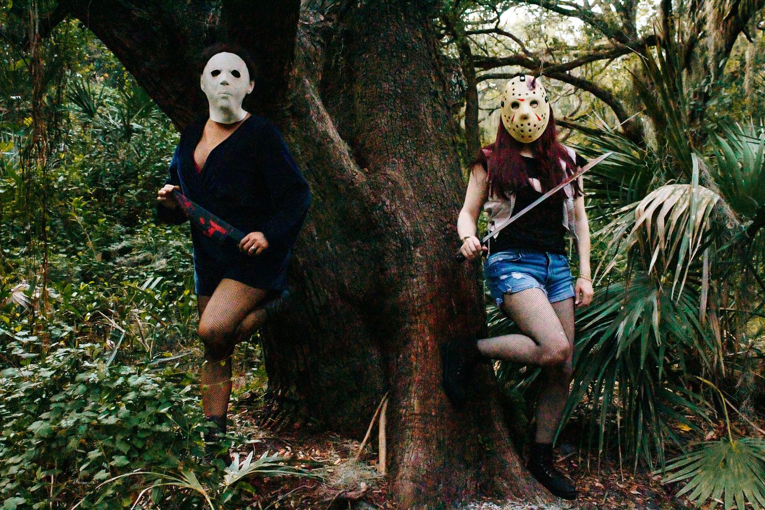 Two masked figures pose dramatically by a large tree trunk in a lush forest setting.