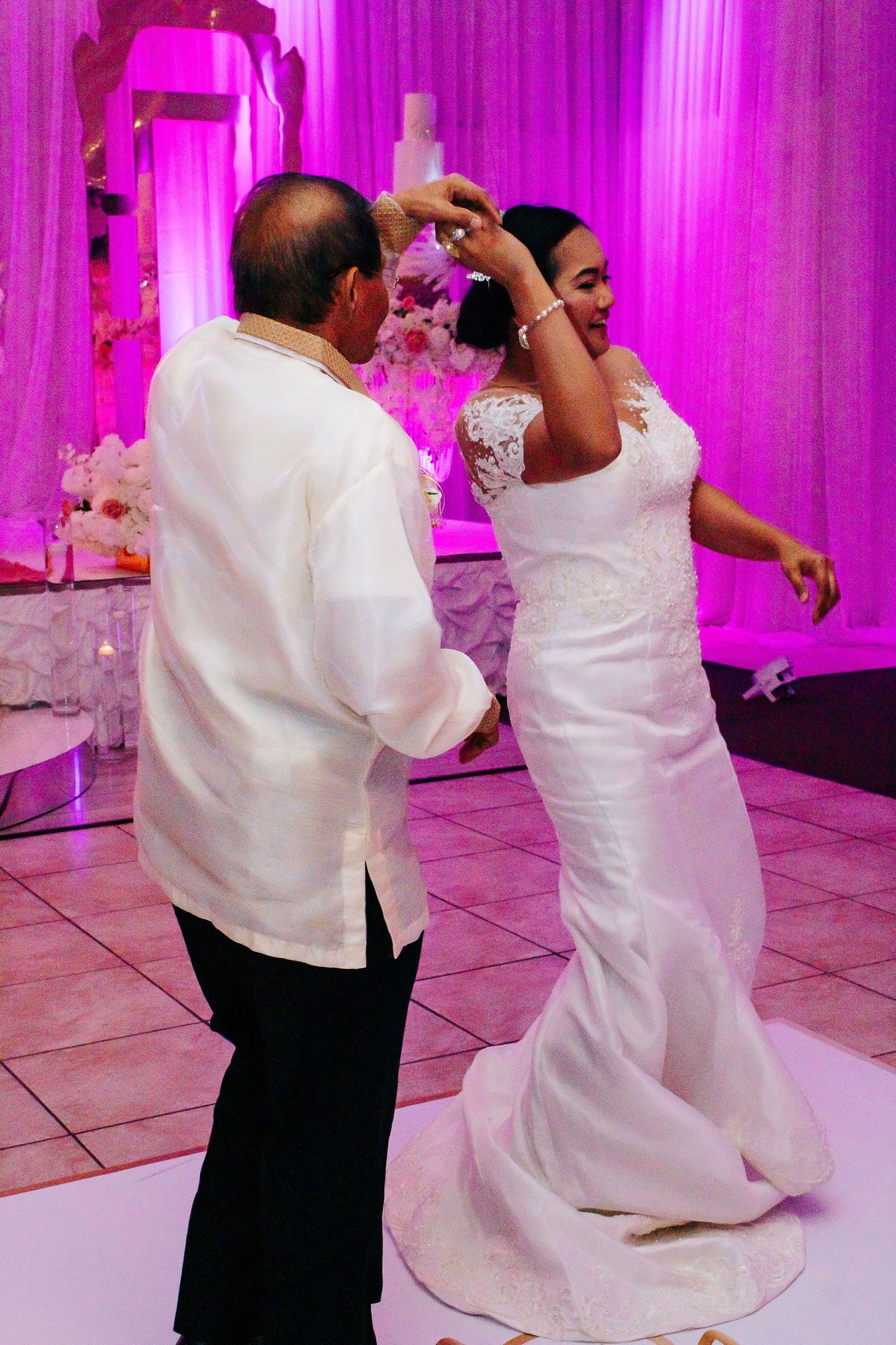 Wedding dance scene with couple in white attire under pink lighting.