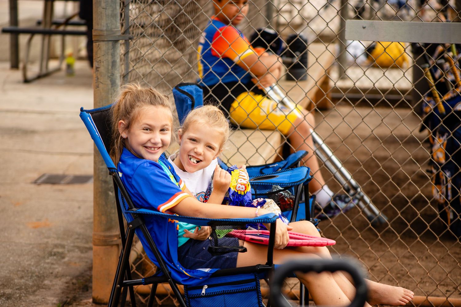 Young spectators enjoy watching baseball game from folding chairs near dugout fence.