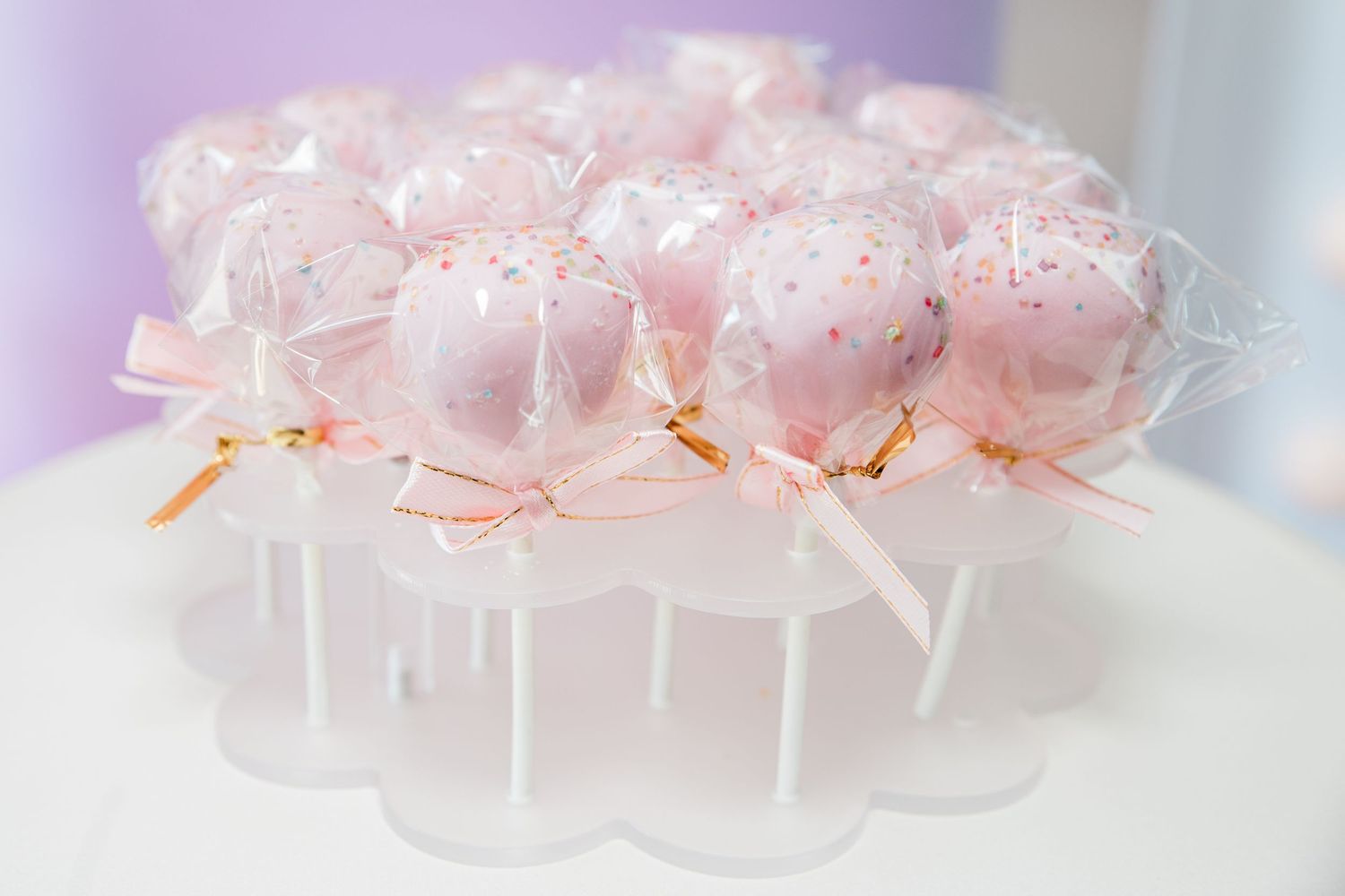Pink cake pops wrapped in clear cellophane with pink ribbon bows displayed on a white acrylic stand.