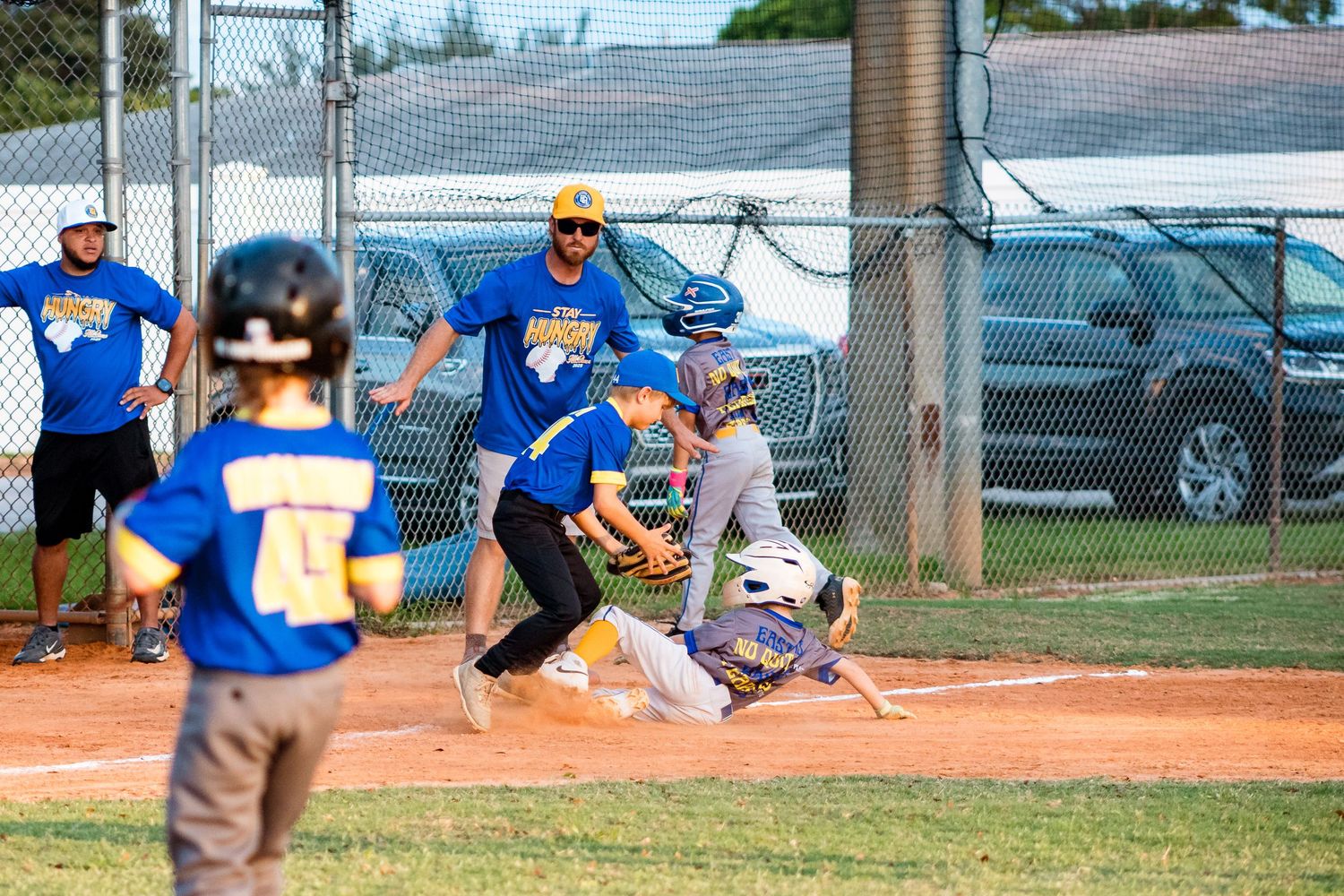 Young baseball players wearing blue and white uniforms compete during a youth baseball game on a dirt diamond field.