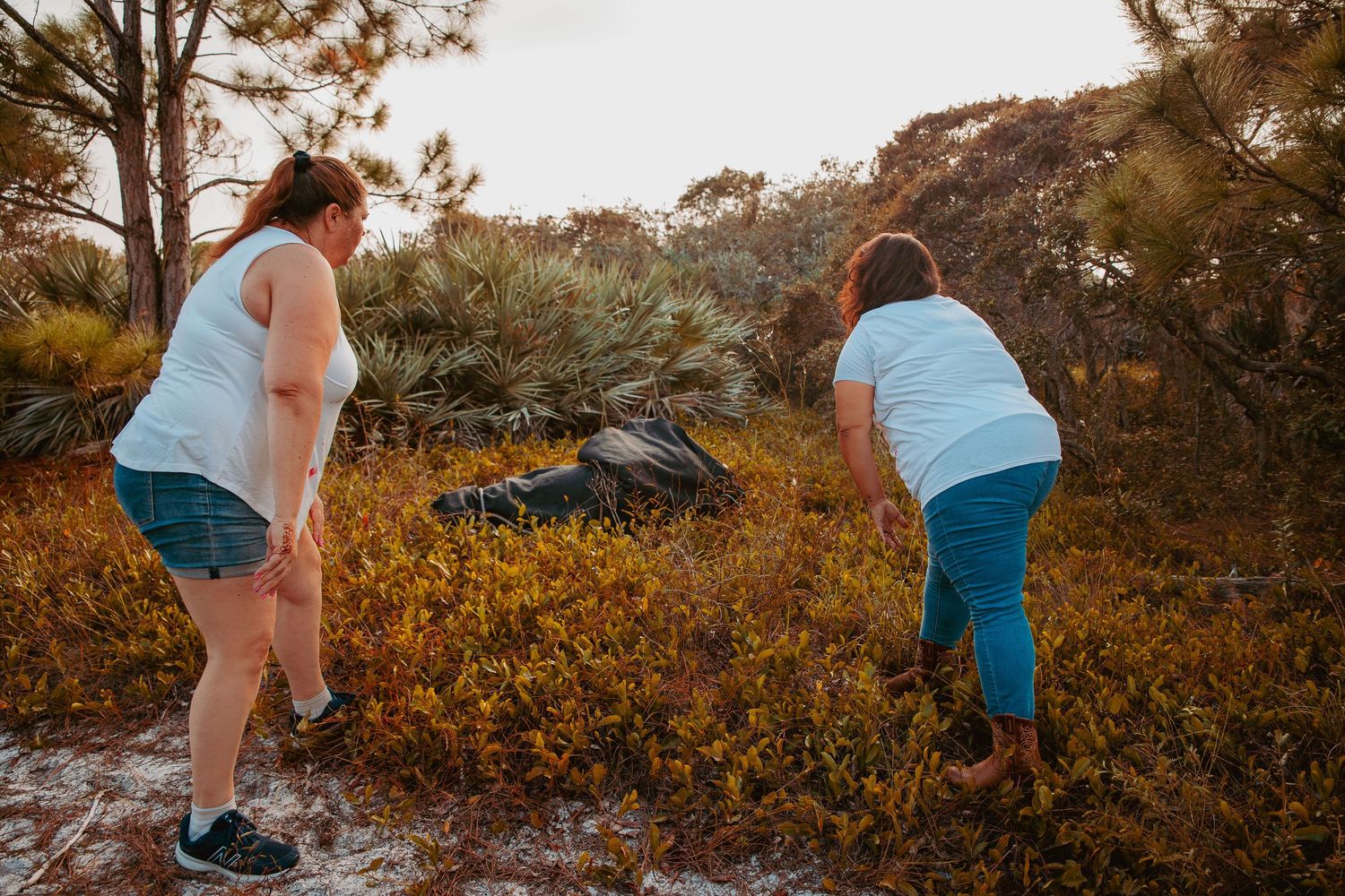 A person in shorts bends down to pick wildflowers in a golden-lit meadow at sunset.
