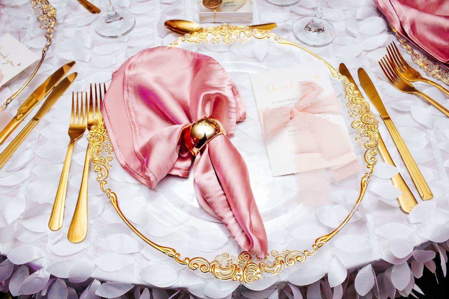 Elegant place setting with pink napkin, gold cutlery, and decorative charger plate on white tablecloth.