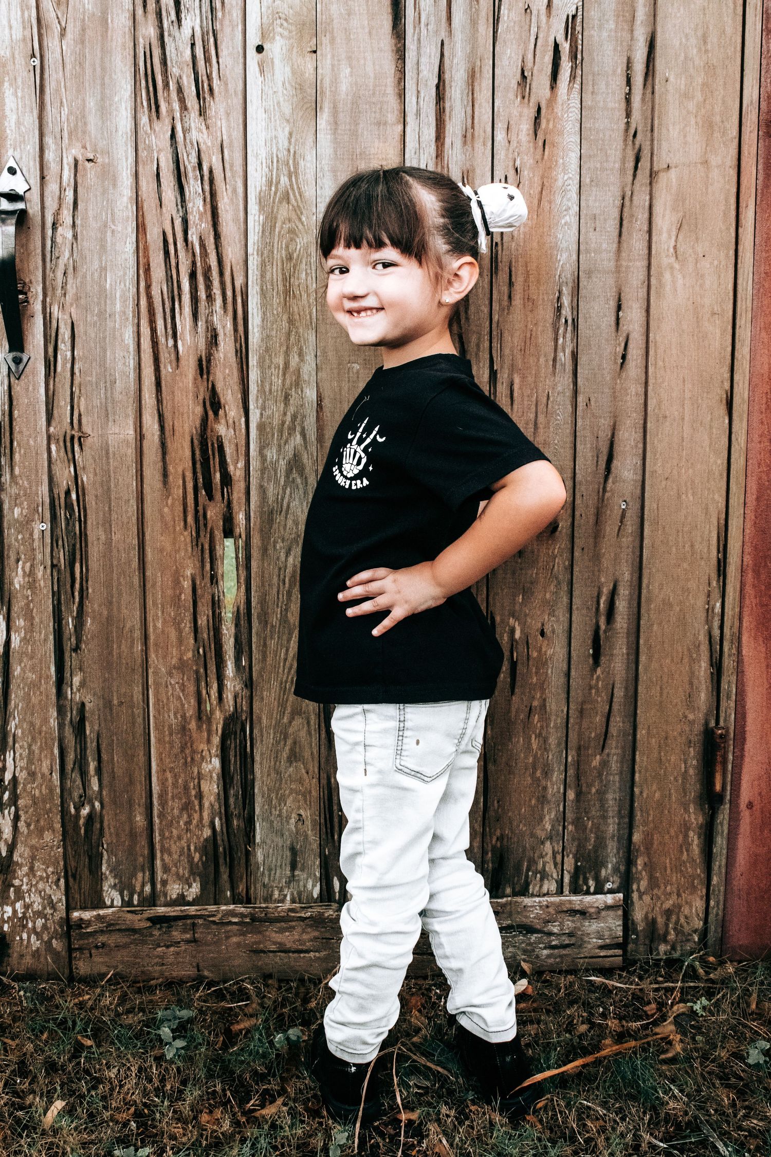 Child in black shirt and white pants smiling while posing against weathered wood background.