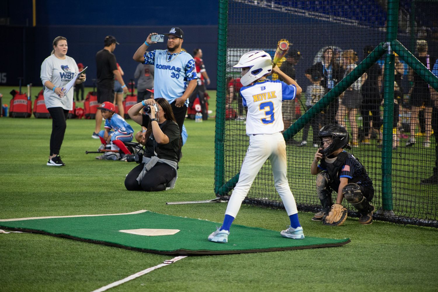 Baseball practice session with multiple players and photographers in batting cage area.