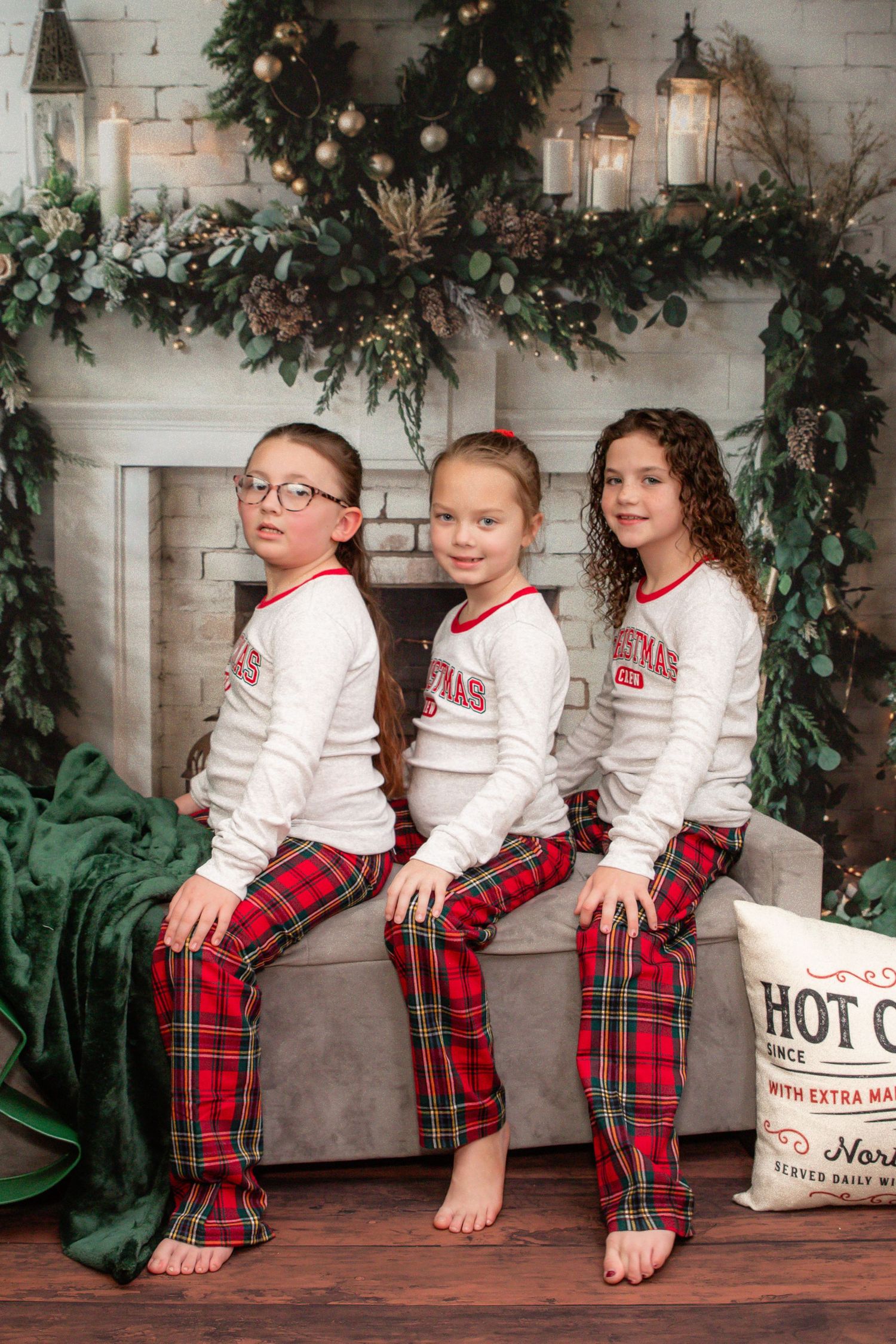Three children in matching plaid pajamas sit on wooden floor with Christmas decor and garland in background.