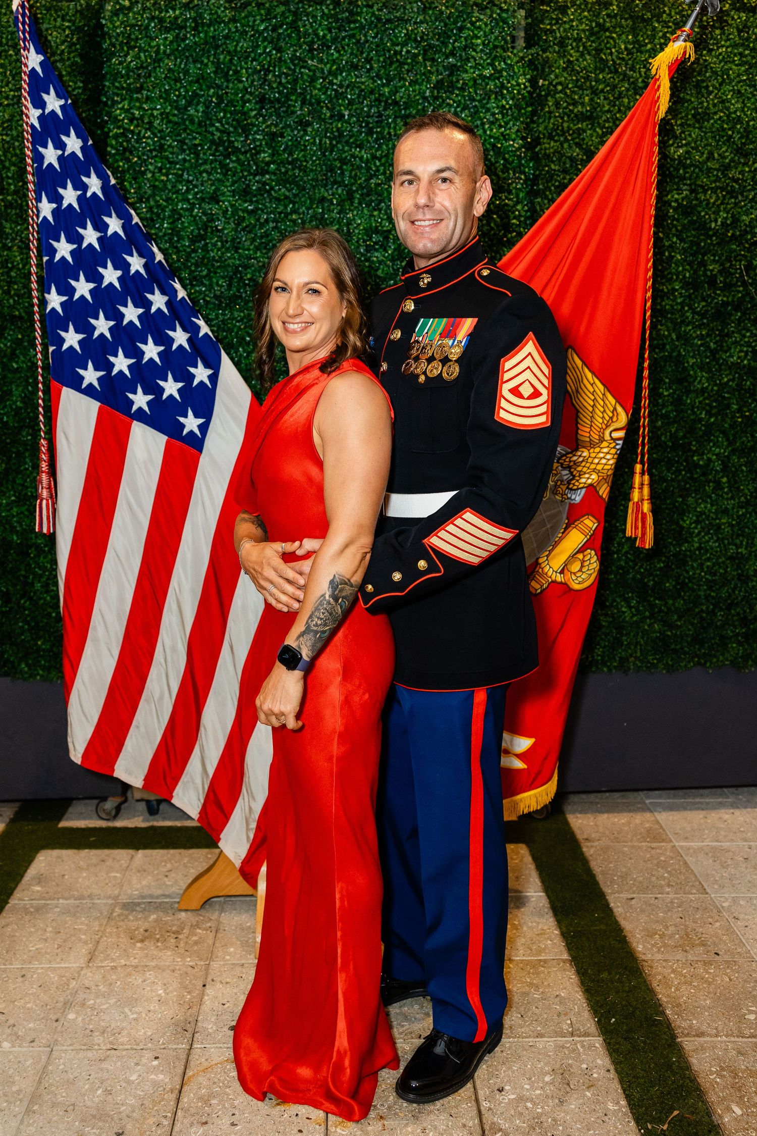 Military service members pose with American and Marine Corps flags against a green hedge backdrop at a formal event.
